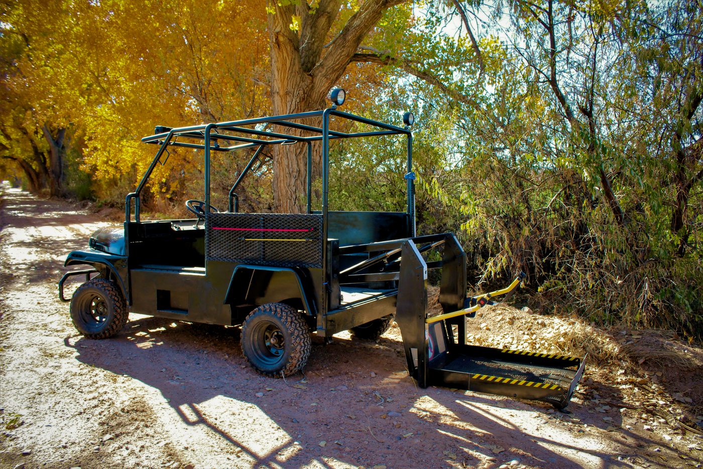 The black Buddy Buggy parked on a dirt trail under cottonwoods in fall color, rear wheelchair lift lowered