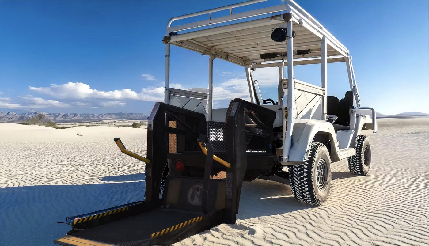 The white Buddy Buggy parked on sand dunes with its rear wheelchair lift lowered to the ground